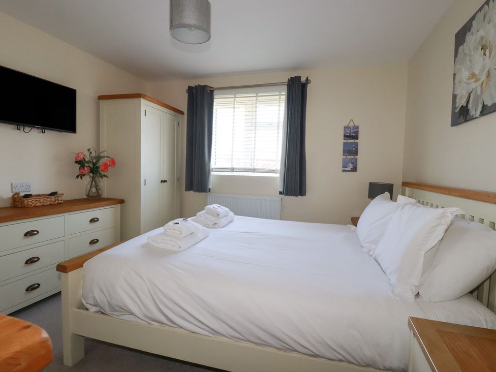 A bedroom with a bed and folded towels a window with dark curtains a dresser with a vase of flowers a wardrobe and a wall-mounted TV at Lodge 18 in St Merryn