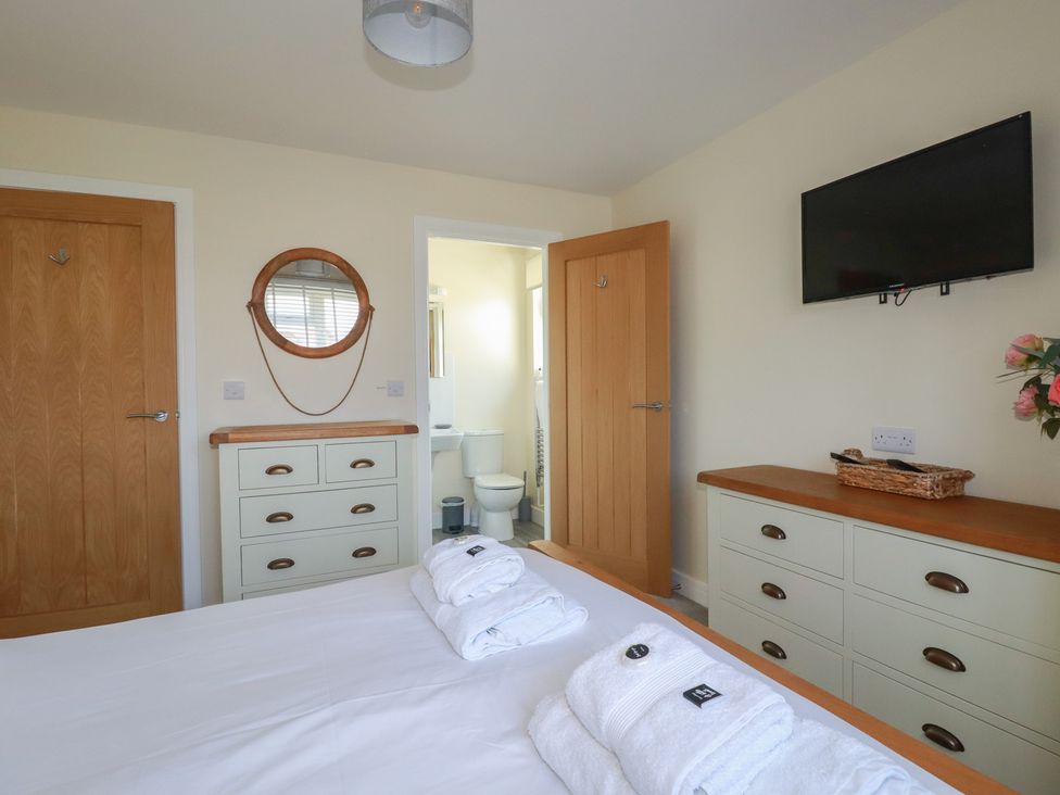 A bedroom with white bed linens and folded towels on the bed a dresser with drawers a wall-mounted TV and an open door leading to a bathroom at Lodge 18 in St Merryn