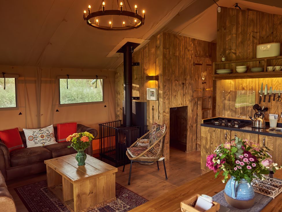 An interior of a wooden cabin with a sofa, wooden coffee table, wicker chair, wood-burning stove, and a kitchen area with a gas stove and flowers in vases at Bramley Lodge in Coxbridge near Glastonbury