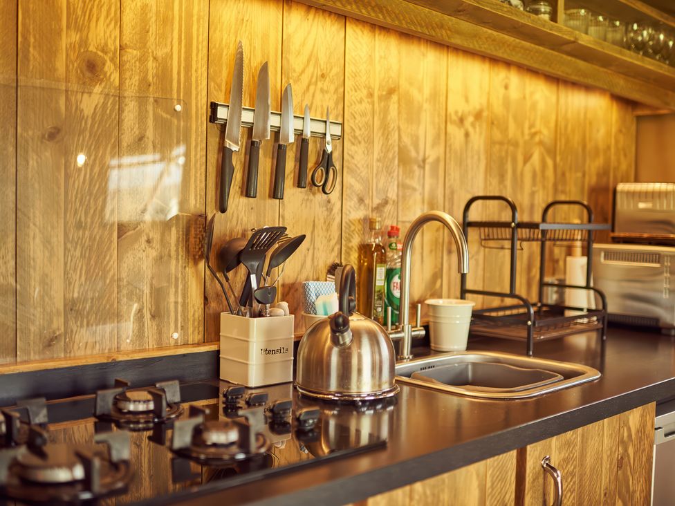 A kitchen counter with a stovetop, utensils holder, knives on a magnetic strip, a kettle, a sink, dish soap, and a dish drying rack at Bramley Lodge in Coxbridge near Glastonbury