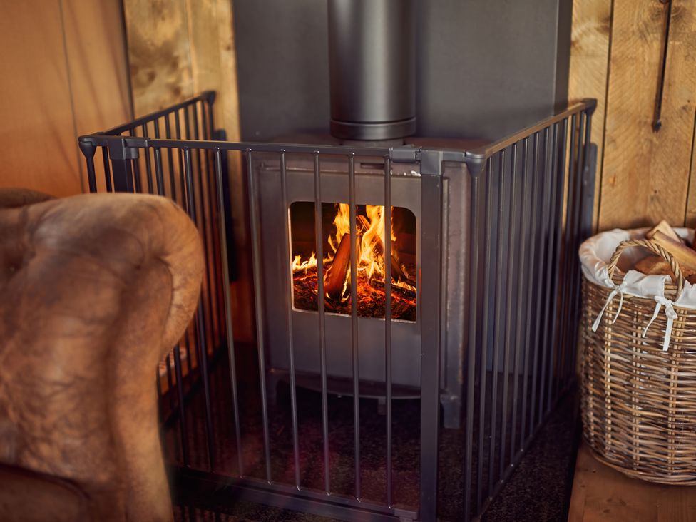 A wood stove with fire inside surrounded by a safety gate next to a leather chair and a basket of firewood at Bramley Lodge in Coxbridge near Glastonbury