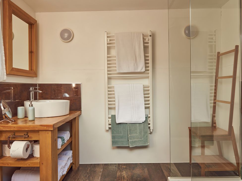 A bathroom with a wooden vanity unit with a white round sink and mirror a towel radiator with towels and a wooden chair behind a glass shower door at Bramley Lodge in Coxbridge near Glastonbury