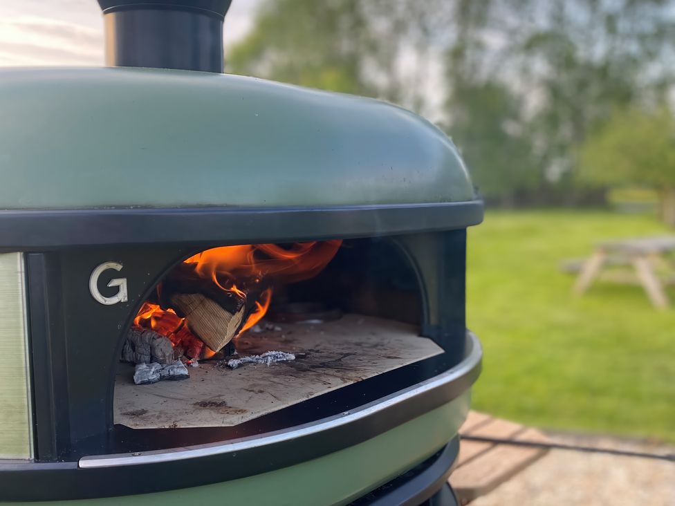 An outdoor wood-fired oven with a burning fire inside in a garden with trees and a wooden picnic table
