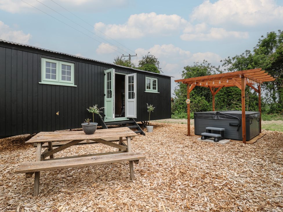 An outdoor area with a wooden picnic table a black caravan with mint green windows and doors and a hot tub with wooden pergola at The Heron in Lode near Bottisham