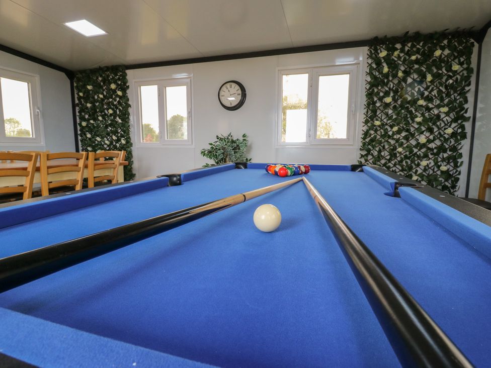 A billiards room with a blue pool table two pool cues a rack of balls a clock and six wooden chairs at The Heron in Lode near Bottisham