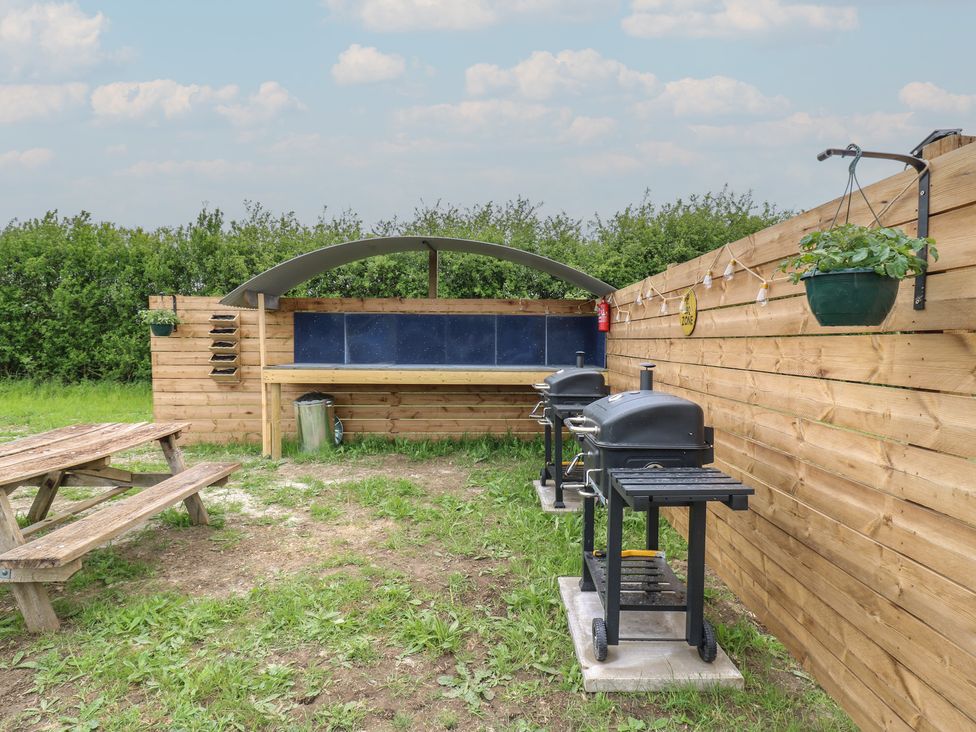 An outdoor area with two black barbecues a wooden picnic table a covered wooden bench and green plants at The Heron in Lode near Bottisham