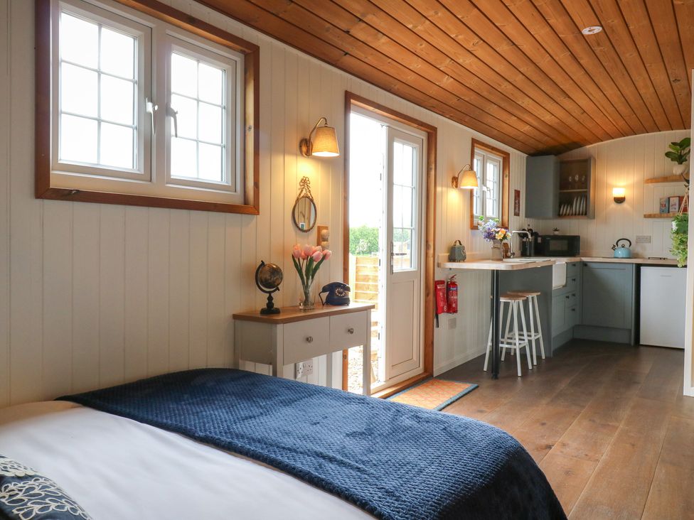 A small room with a bed a side table with flowers and a vintage phone an open door leading outside and a kitchenette with a countertop and stools at Barn Owl in Lode near Bottisham