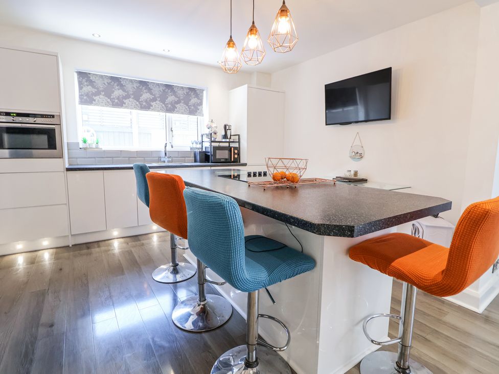 A kitchen with a black countertop island, blue and orange bar stools, pendant lights, an oven, and a wall-mounted TV at 67 Pendre Avenue in Prestatyn