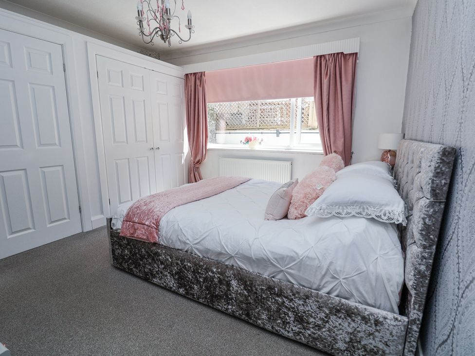 A bedroom with a bed with pillows and blankets a window with curtains a chandelier and built in white wardrobes at 67 Pendre Avenue in Prestatyn