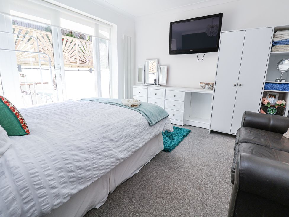 A bedroom with a bed and white bedding a wall-mounted TV a white dresser a white wardrobe and a black armchair at 67 Pendre Avenue in Prestatyn
