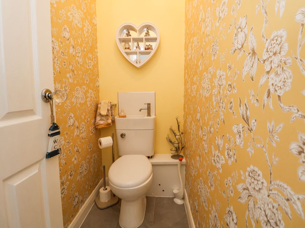 A small toilet room with a toilet and sink, floral wallpaper, a heart-shaped wall shelf, and a door with a knob and WC sign at 67 Pendre Avenue in Prestatyn