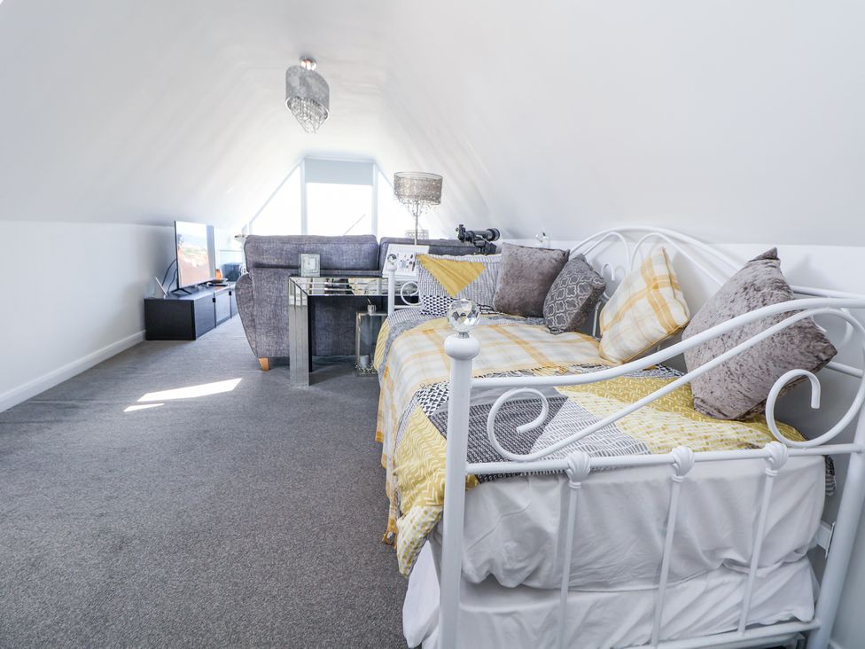 An attic room with a white metal bed, grey sofa, glass side table, television on a black stand and a window at 67 Pendre Avenue Prestatyn
