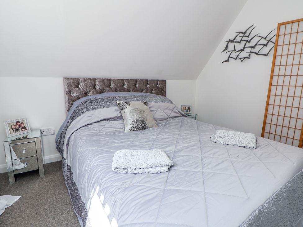 A bedroom with a bed covered in gray bedding two towels on the bed mirrored bedside tables family photos and wall art at 67 Pendre Avenue in Prestatyn