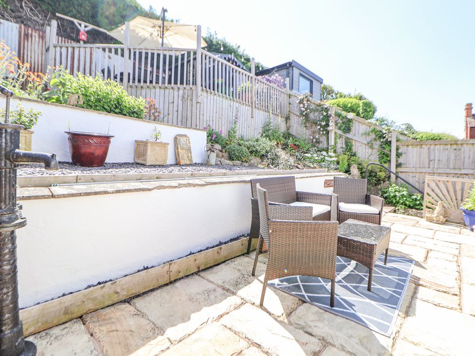 A patio area with wicker furniture and a small table on a rug surrounded by plants and a wooden fence at 67 Pendre Avenue in Prestatyn