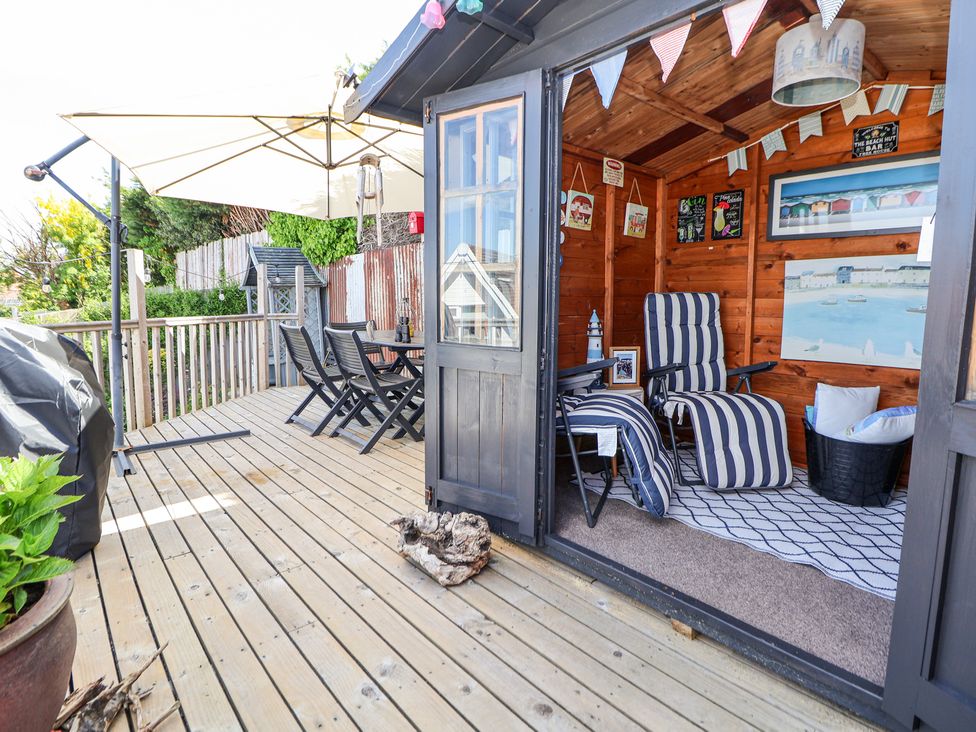 An outdoor wooden deck with a dining table and chairs a large umbrella and a wooden shed with striped lounge chairs and decorations at 67 Pendre Avenue in Prestatyn