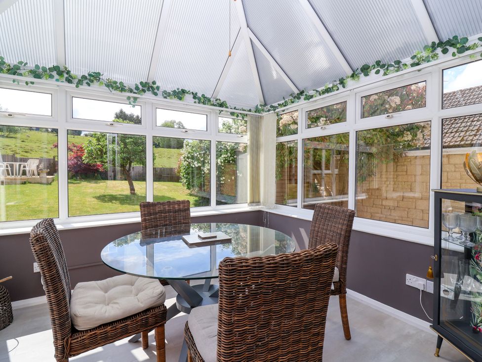 A conservatory with a round glass table and wicker chairs overlooking a garden at Hillside in Bishops Cleeve