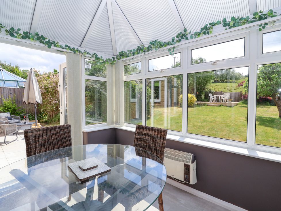A conservatory with a glass dining table wicker chairs and a view of a garden at Hillside in Bishops Cleeve