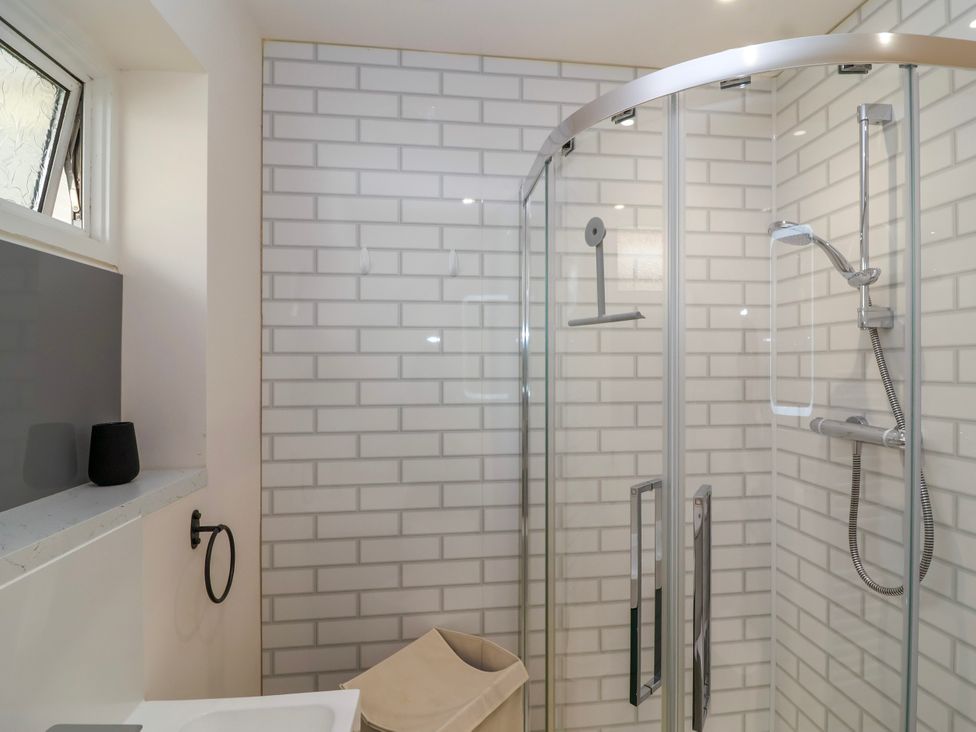A bathroom with a glass shower enclosure white brick wall tiles a window and a laundry basket at Hillside in Bishops Cleeve