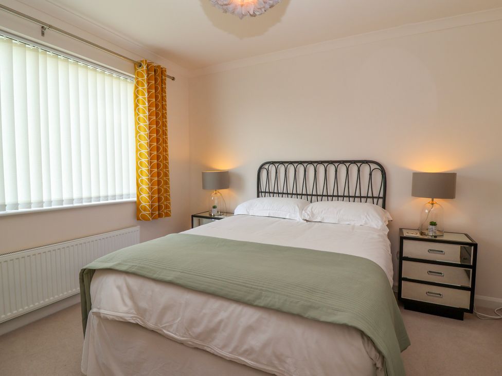 A bedroom with a double bed with green throw and white bedding two bedside tables with lamps and a window with yellow curtains at Hillside in Bishops Cleeve
