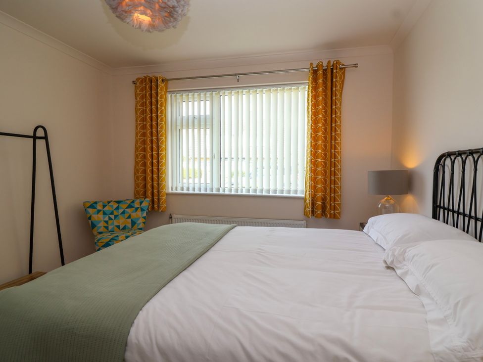 A bedroom with a bed against the wall a window with yellow curtains a chair with a geometric pattern and a lamp at Hillside in Bishops Cleeve