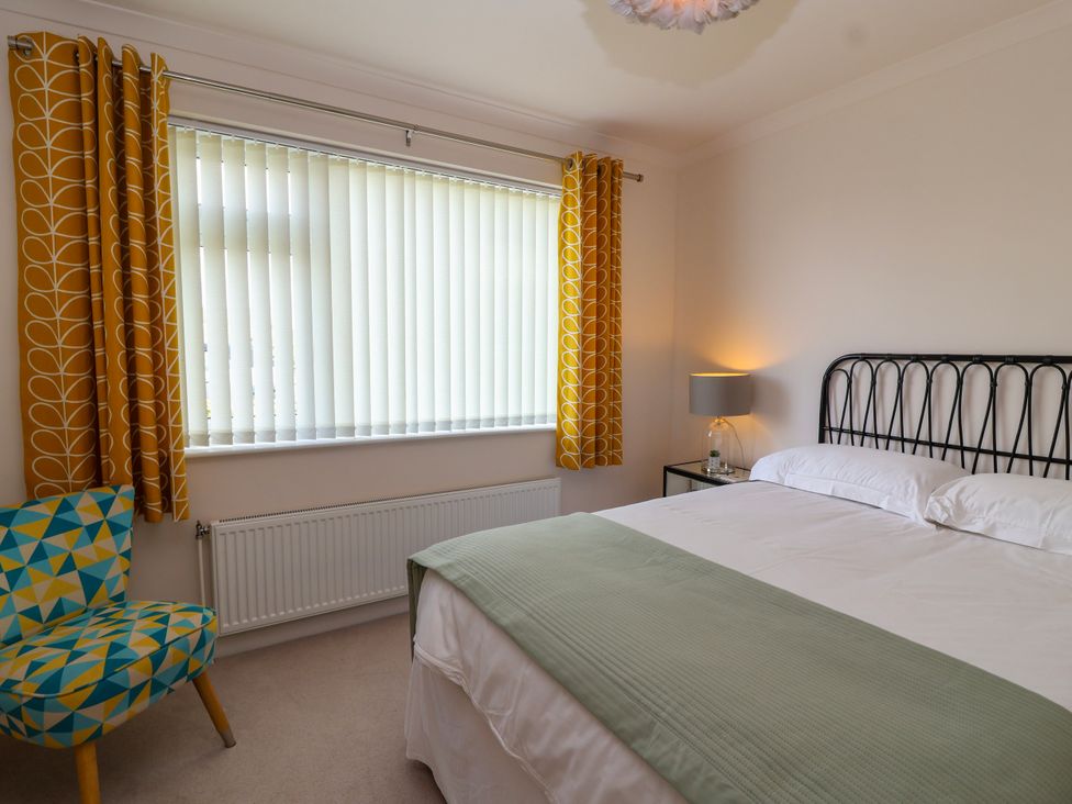 A bedroom with a double bed, a bedside table with a lamp, a patterned chair, and a window with vertical blinds and yellow curtains at Hillside in Bishops Cleeve