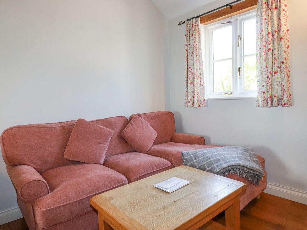 A pink sofa with two cushions a wooden coffee table and a window with floral curtains at Rose's Stable in Bodrean near Truro