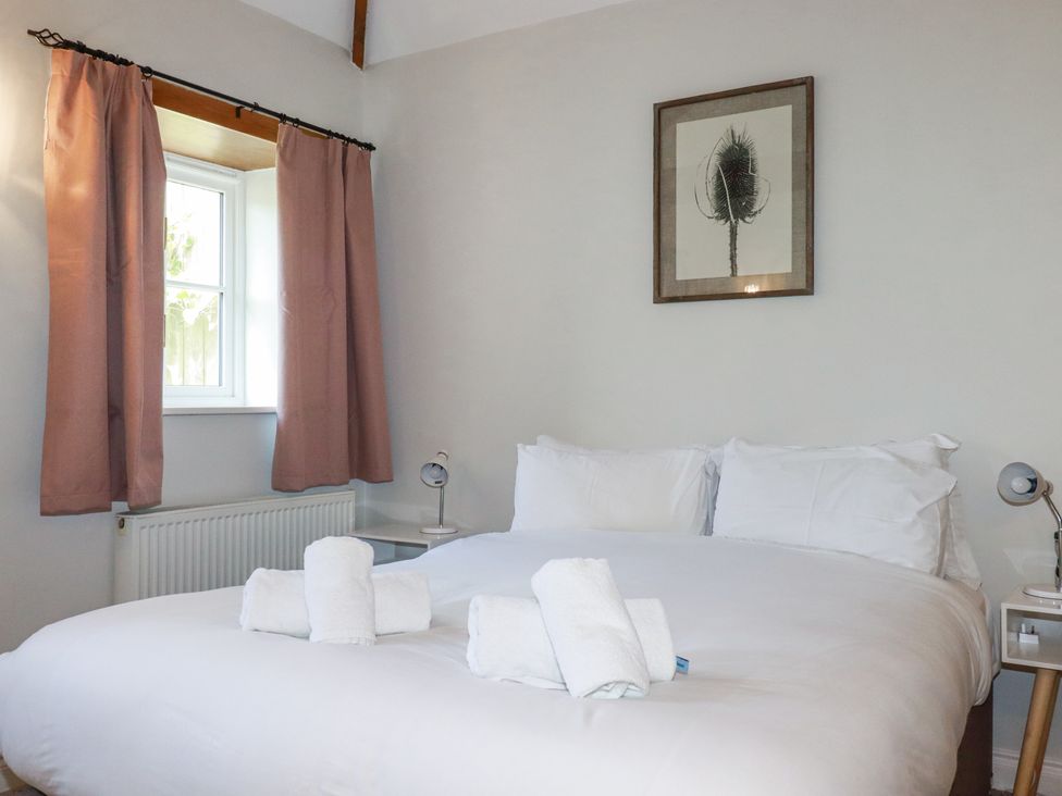 A bedroom with a double bed with white linens and towels a window with pink curtains and framed artwork on the wall at Rose's Stable in Bodrean near Truro