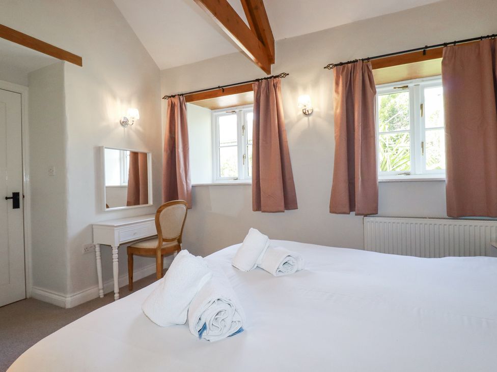 A bedroom with two windows with curtains a white dressing table with a chair and rolled towels on a bed at Rose's Stable in Bodrean near Truro