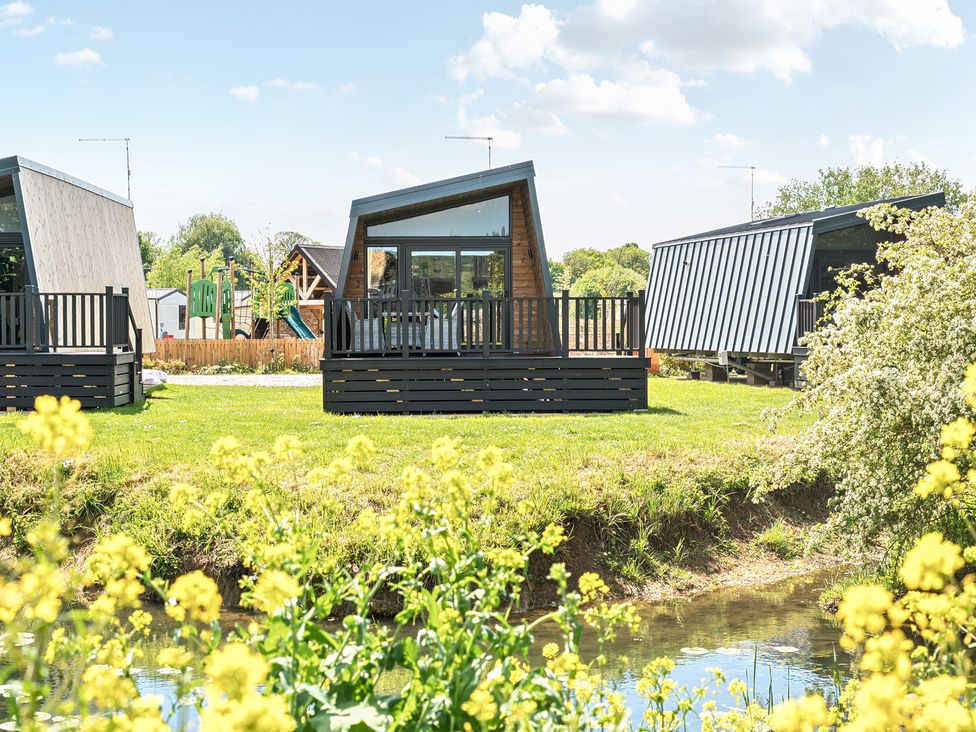A grassy area with modern small cabins next to a small pond with yellow flowers and a playground in the background at Celestial Skylight - Billing Aquadrome in Billing Aquadrome Holiday Park