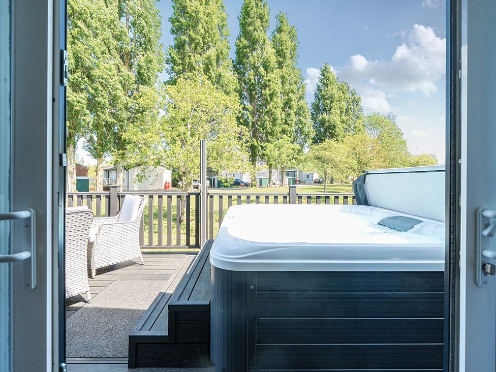 A patio with a hot tub and two wicker chairs on a deck outside near trees at Lakeside Lullaby - Billing Aquadrome Holiday Park
