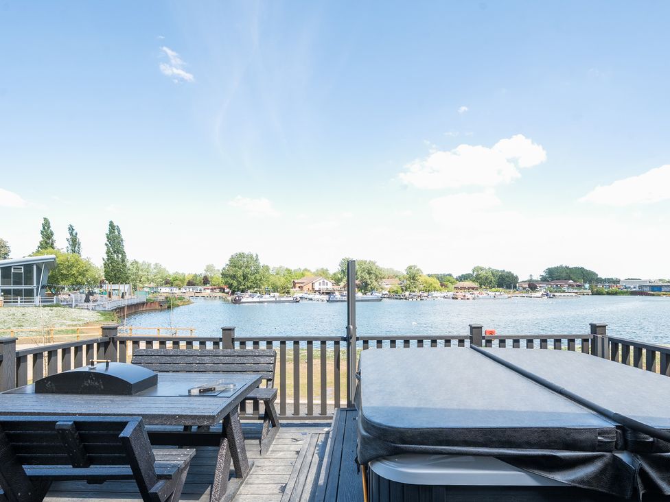 A deck with wooden table and benches and a covered hot tub overlooking a lake at Skylight Stargazer - Billing Aquadrome Holiday Park