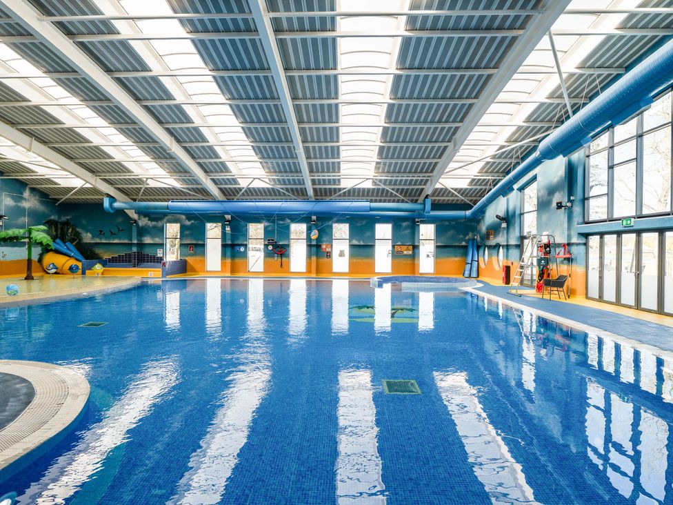 An indoor swimming pool with large windows ceiling lights a water slide and lifeguard chair at Brookside Bliss - Billing Aquadrome in Billing Aquadrome Holiday Park