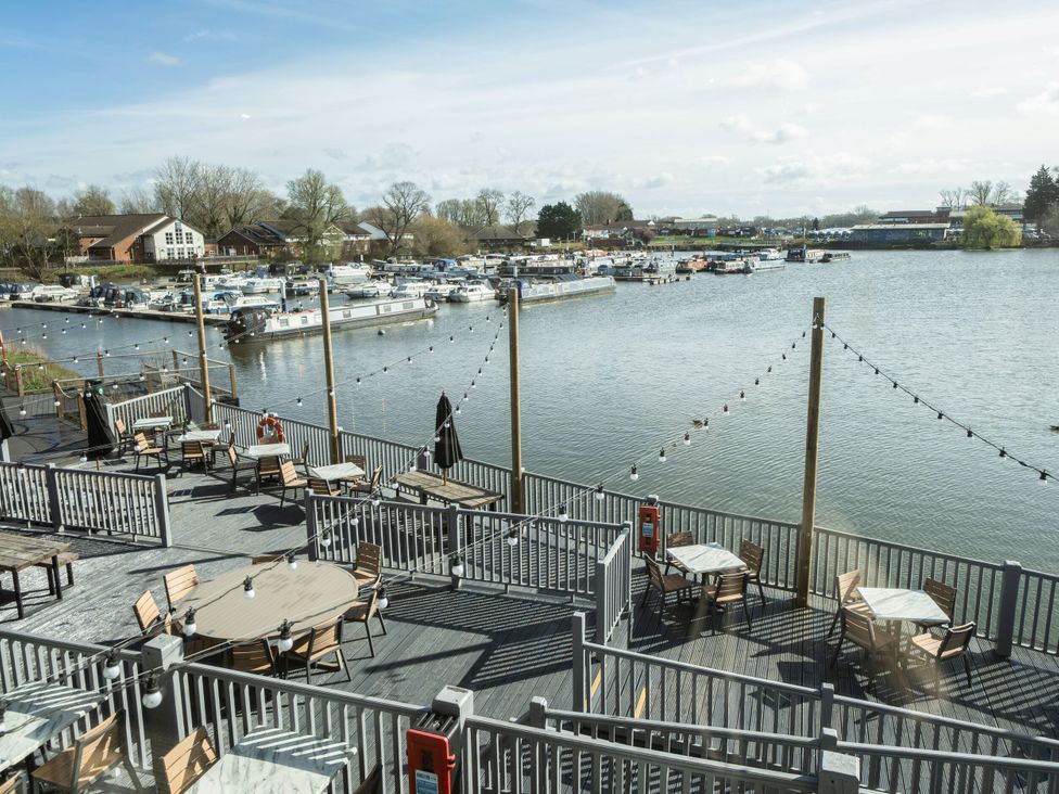 An outdoor dining area with tables and chairs on a deck overlooking a marina with boats and houses in the background at Brookside Bliss - Billing Aquadrome