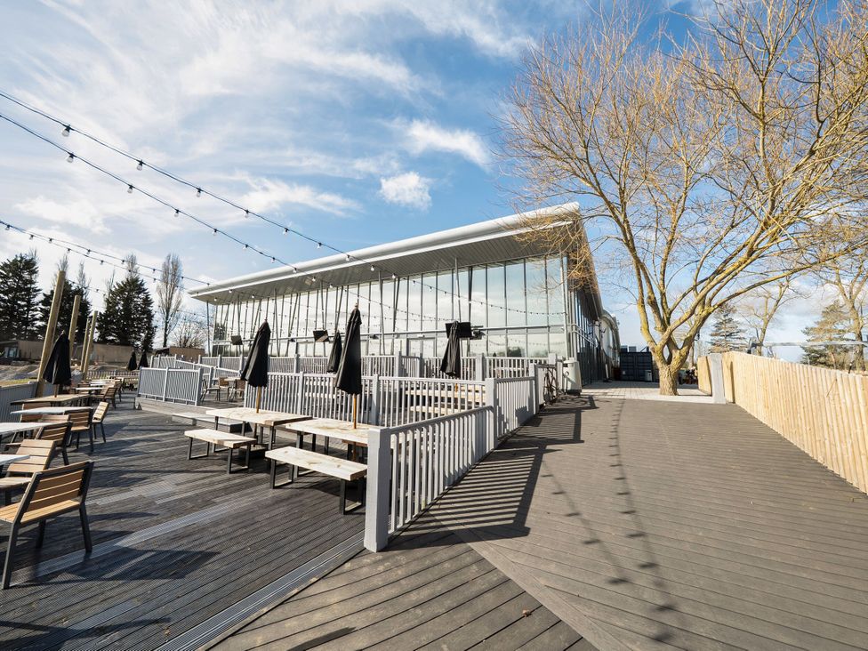 An outdoor seating area with wooden tables and benches under string lights beside a glass building and trees at Brookside Bliss - Billing Aquadrome in Billing Aquadrome Holiday Park