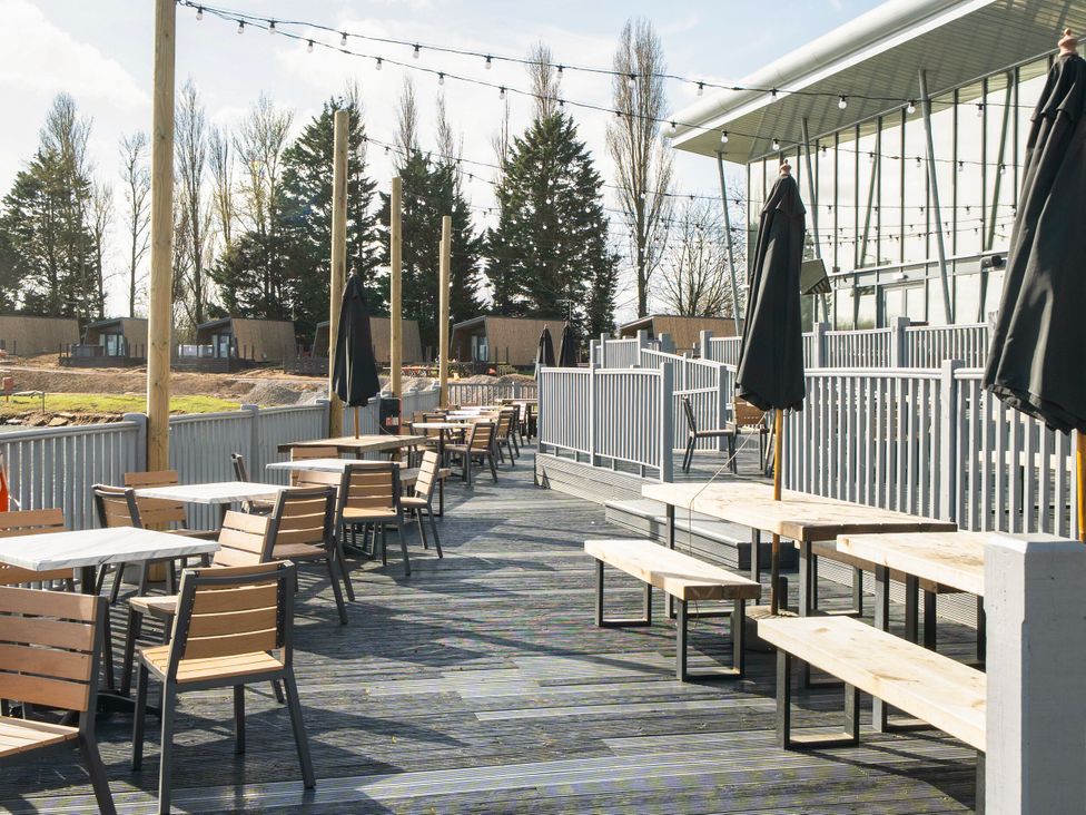An outdoor dining area with tables chairs and umbrellas next to a building with large windows at Brookside Bliss - Billing Aquadrome in Billing Aquadrome Holiday Park