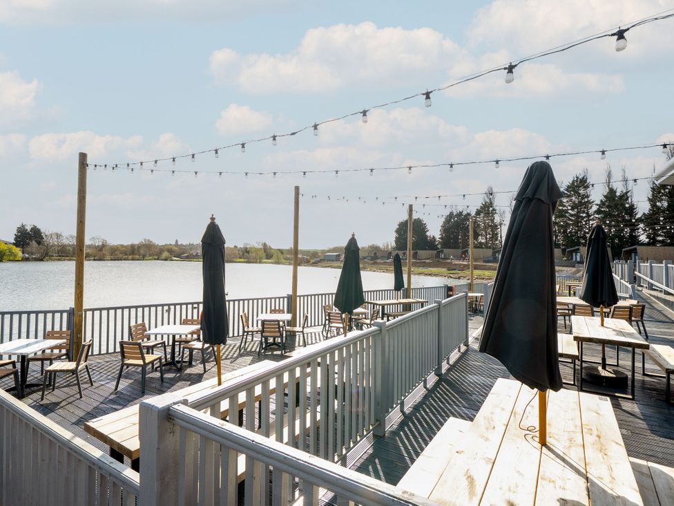 An outdoor deck area with tables chairs and closed umbrellas overlooking a lake with string lights above at Brookside Bliss - Billing Aquadrome
