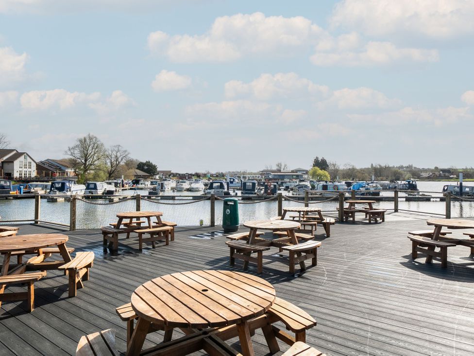 An outdoor deck with wooden picnic tables and benches overlooking a marina with boats at Brookside Bliss - Billing Aquadrome in Billing Aquadrome Holiday Park