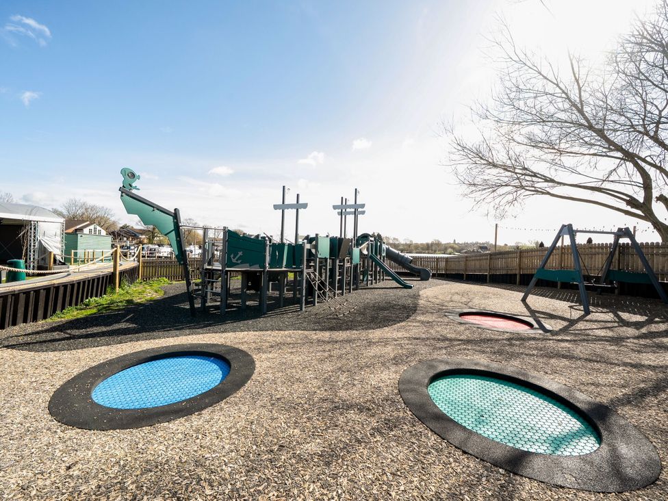 A playground with a ship-themed climbing structure slides swings and round trampolines at Brookside Bliss - Billing Aquadrome in Billing Aquadrome Holiday Park