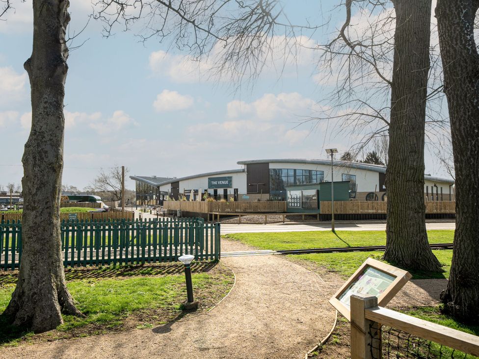 An outdoor scene with a gravel path between trees and green fences facing a modern building labeled The Venue at Billing Aquadrome Holiday Park