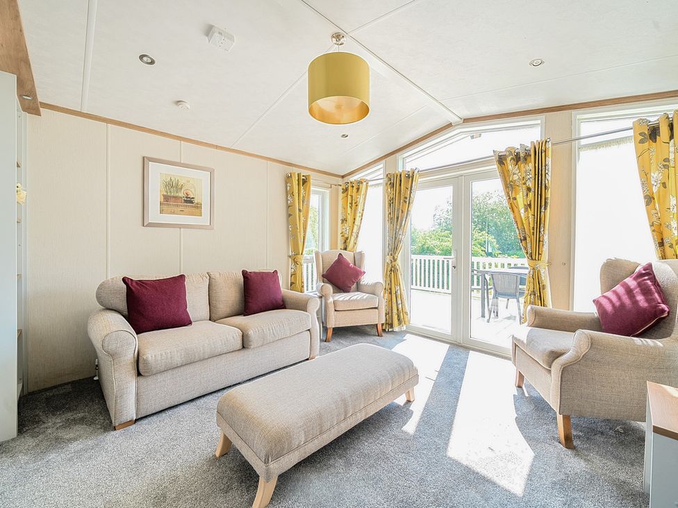 A living room with beige sofa chairs and an ottoman near large glass doors with yellow floral curtains at Seaside Serenity - Hayling Island in Hayling Island Holiday Park