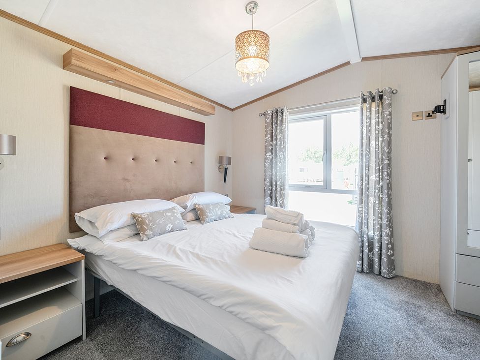 A bedroom with a double bed with rolled towels and floral cushions near a window with curtains at Seaside Serenity - Hayling Island Hayling Island Holiday Park