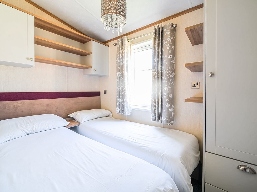 A bedroom with two single beds with white bedding a window with curtains and built-in shelves at Seaside Serenity - Hayling Island Holiday Park