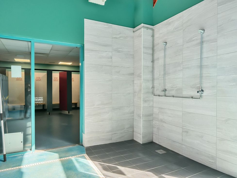 An indoor shower area with two shower heads and tiled walls next to a glass door leading to a changing room with benches at Island Breeze - Hayling Island in Hayling Island Holiday Park