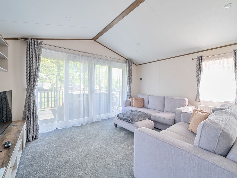 A living room with two sofas a footstool a television and large windows with curtains at Wave Whisperers - Hayling Island in Hayling Island Holiday Park
