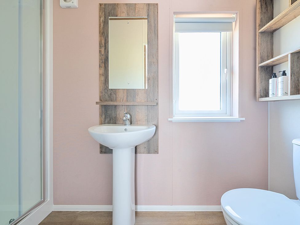 A bathroom with a pedestal sink mirror window toilet and wooden shelves at Wave Whisperers - Hayling Island Hayling Island Holiday Park