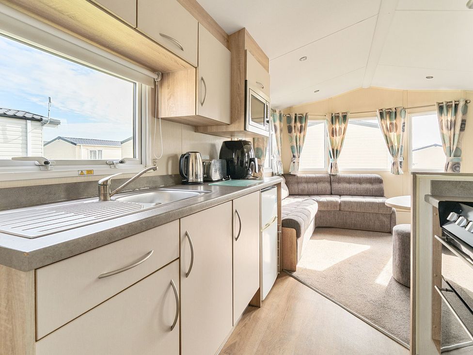 A kitchen and sitting area with a sink, countertop appliances, cabinets, sofa, windows with curtains at The Lookout - Hayling Island in Hayling Island Holiday Park