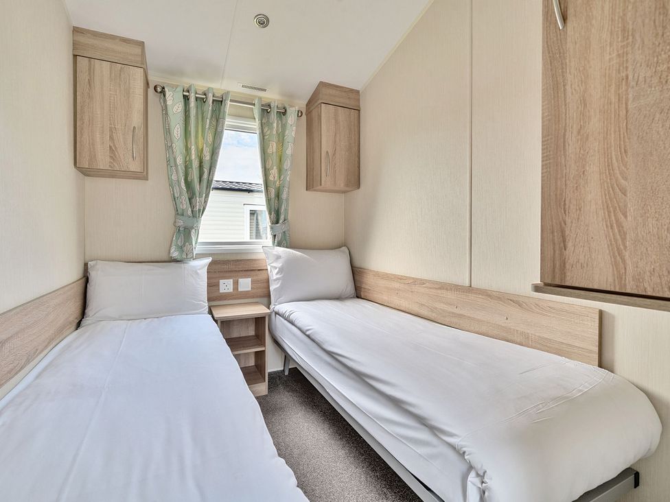 A small bedroom with two single beds separated by a wooden nightstand and a window with patterned curtains at The Lookout - Hayling Island in Hayling Island Holiday Park