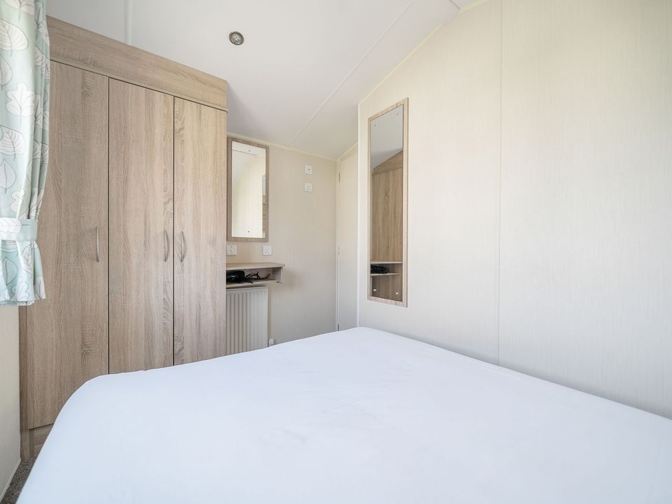 A bedroom with a bed a wooden wardrobe two mirrors and a window with patterned curtains at The Lookout - Hayling Island Hayling Island Holiday Park