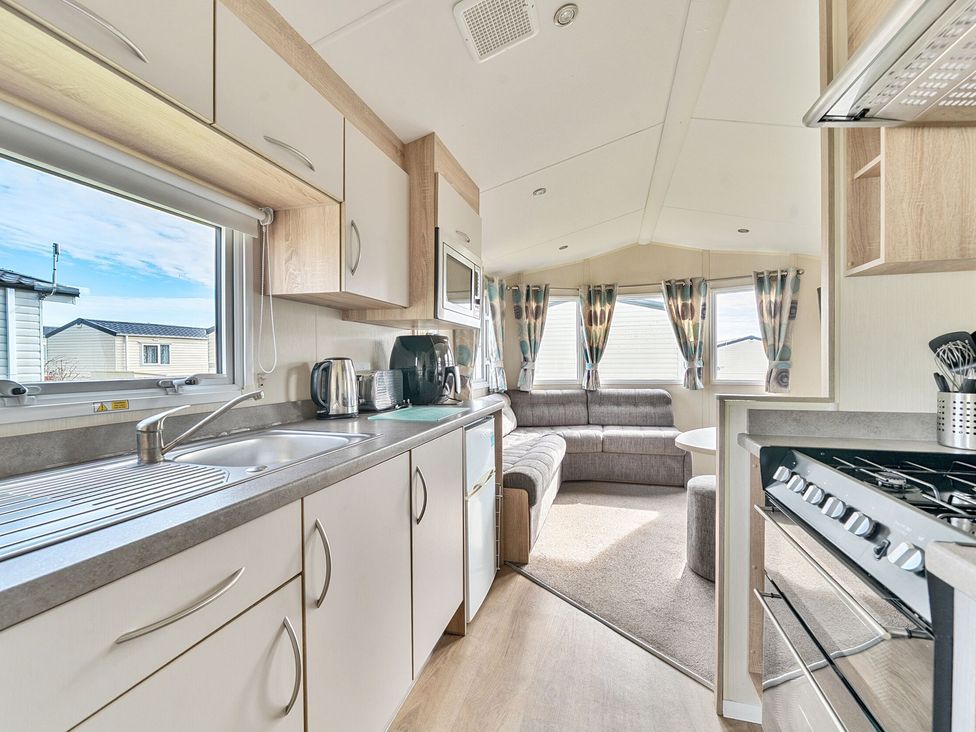 A kitchen with a sink stove and countertop appliances next to a living area with a sectional sofa and windows with curtains at The Lookout - Hayling Island in Hayling Island Holiday Park