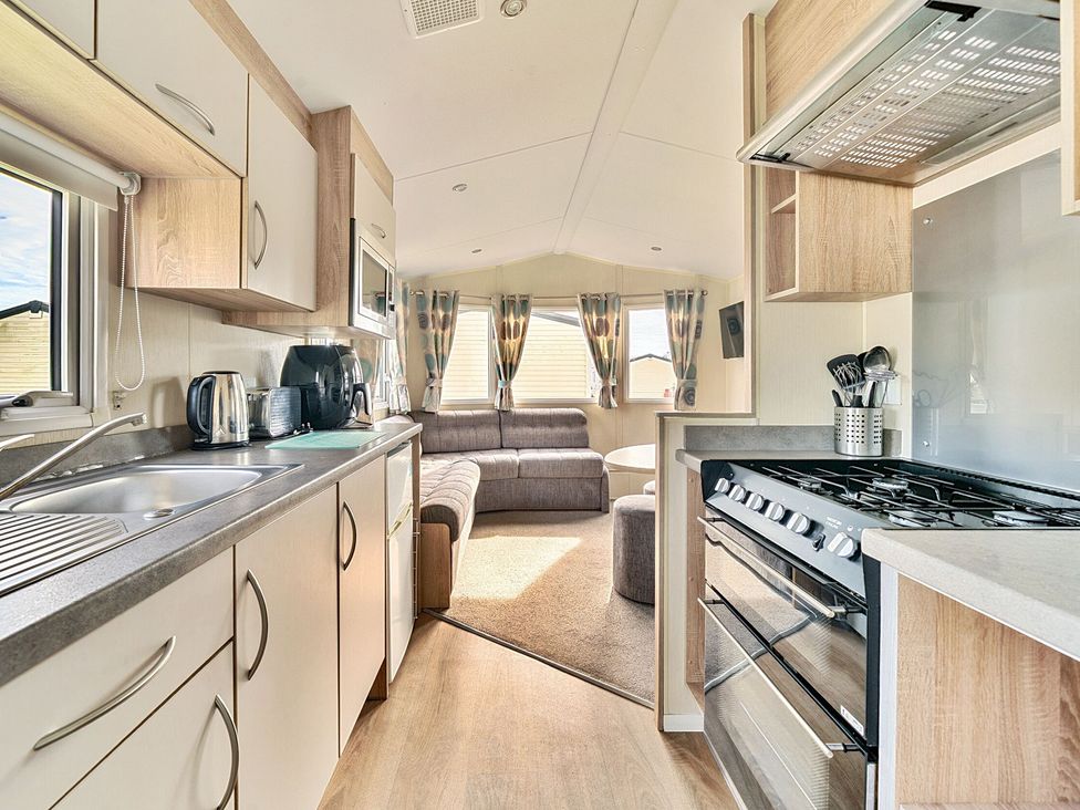 A kitchen with a stove and sink looking into a living room with a sofa and windows at The Lookout - Hayling Island in Hayling Island Holiday Park